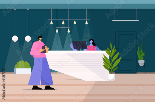 man standing at reception desk and talking to receptionist in hotel or office full length