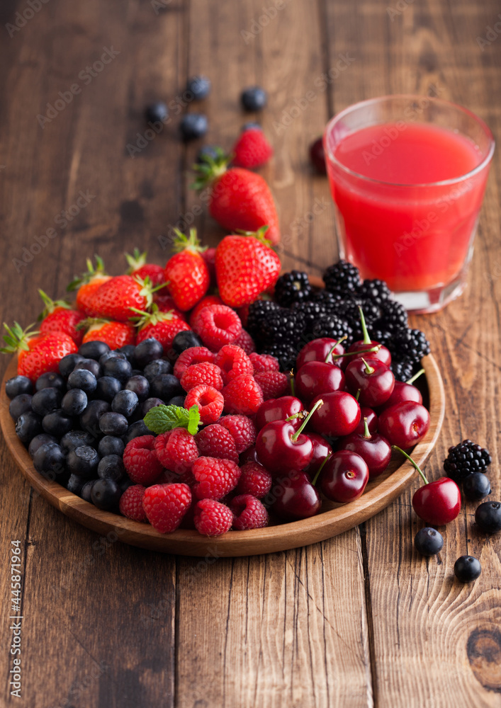 Fresh organic summer berries mix in round wooden tray with glass of juice on light wooden table background. Raspberries, strawberries, blueberries, blackberries and cherries. Fresh organic summer berries mix in round wooden tray with glass of juice on light wooden table background. Raspberries, strawberries, blueberries, blackberries and cherries.