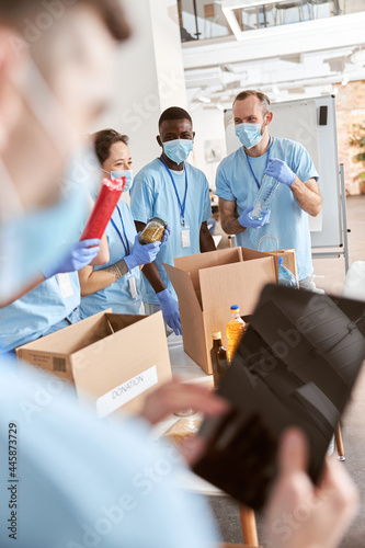 Cheerful volunteers in blue uniform, protective masks and gloves sorting, packing food stuff in cardboard boxes, working together on donation project