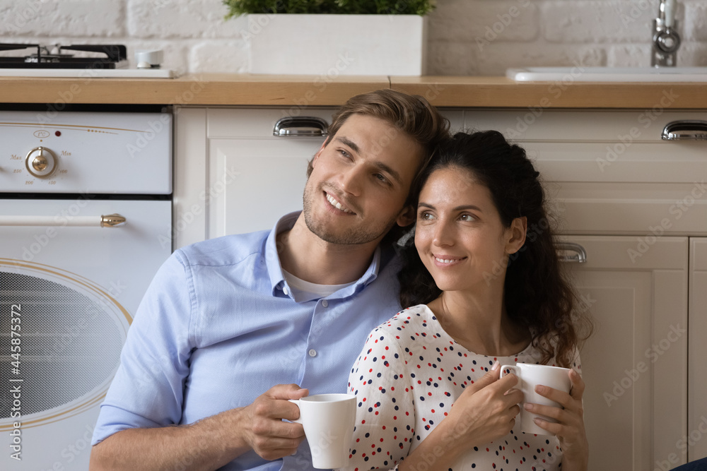 Bonding young married couple sit on floor at kitchen cuddle dream together hold cups with coffee tea. Smiling loving millennial spouses plan future life look at distance enjoy hot drinks at cozy home Bonding young married couple sit on floor at kitchen cuddle dream together hold cups with coffee tea. Smiling loving millennial spouses plan future life look at distance enjoy hot drinks at cozy home
