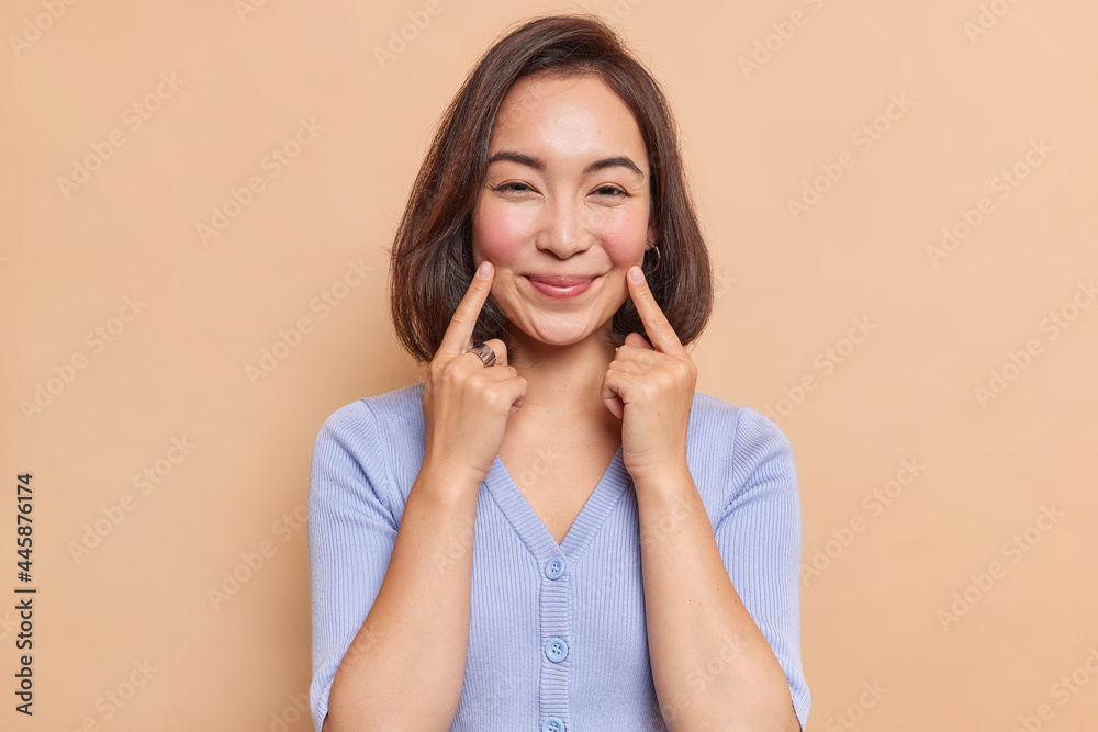 Portrait of cute dark haired woman points at lip corners smiles gently has healthy skin rouge cheeks being in good mood wears blue jumper isolated over beige background. Happy face expressions Portrait of cute dark haired woman points at lip corners smiles gently has healthy skin rouge cheeks being in good mood wears blue jumper isolated over beige background. Happy face expressions