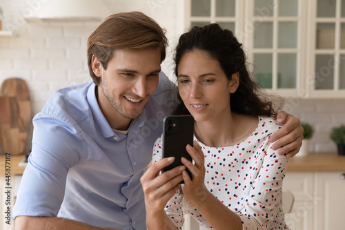 Loving millennial wife show beloved husband consumer goods on phone screen ask opinion discuss online purchases. Happy young couple choose trip at travel agency website plan future vacation together