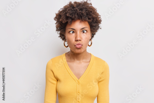 Photo of funny curly haired pretty Afro American woman keeps lips folded crosses eyes makes grimace at camera foolishes around wears yellow jumper isolated over white background has fun alone