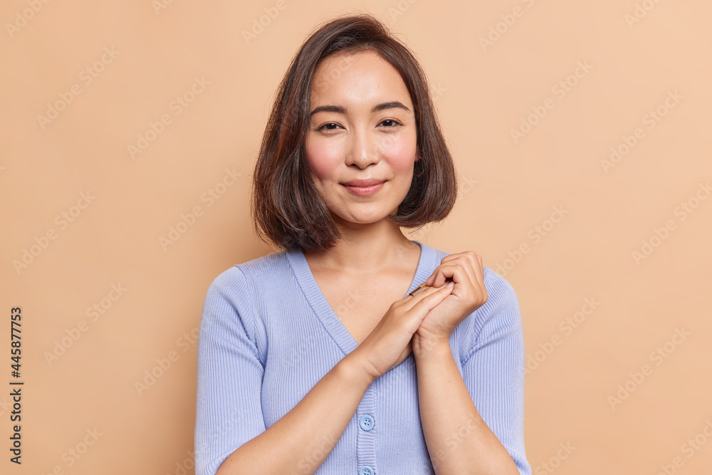 © WHstudio Leushin N - Portrait of good looking brunette young Asian woman has pleased expression healthy skin natural beauty keeps hands together looks mysteriously at camera dressed in blue jumper poses indoor alone