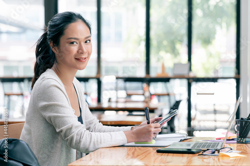 Portrait of smilely casual woman holding smartphone and pen while sitting at the table in office. photo