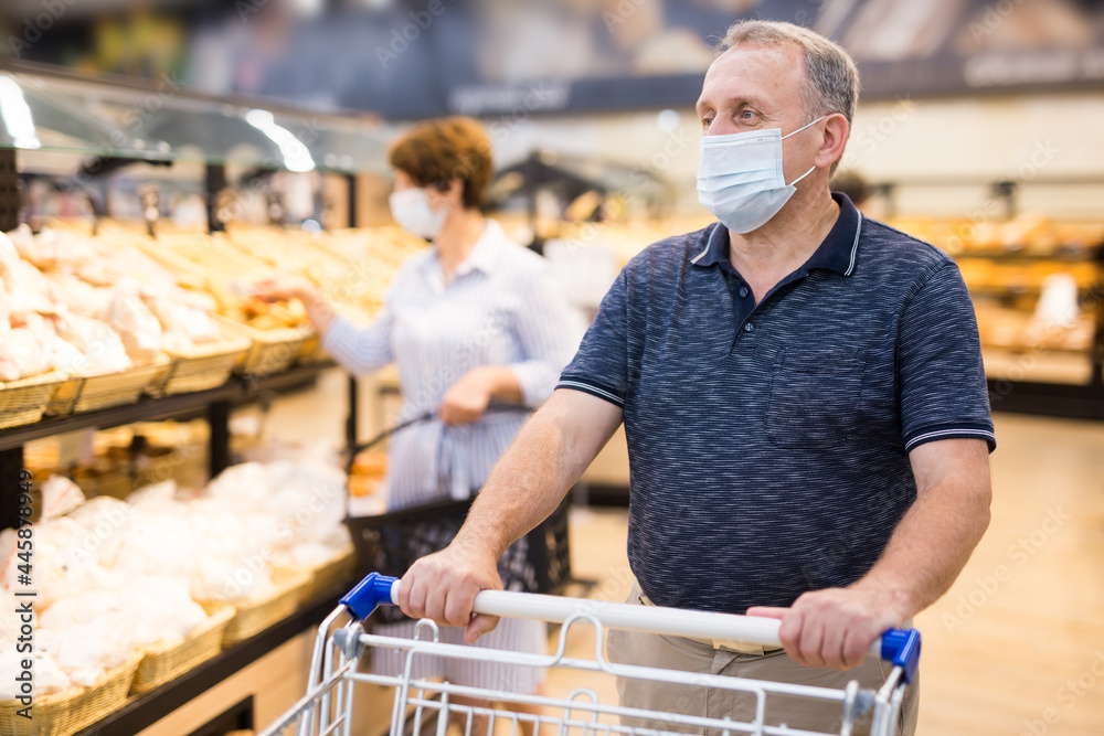 Senior man in protective mask with shopping trolley choosing pastries and products in supermarket