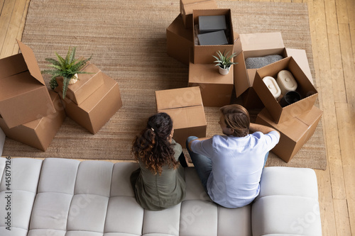 New home owners. Top view of young married couple sit on sofa look at moving boxes with belongings discuss house furnishing. Young man woman spouses roommates plan unpacking things after relocation photo