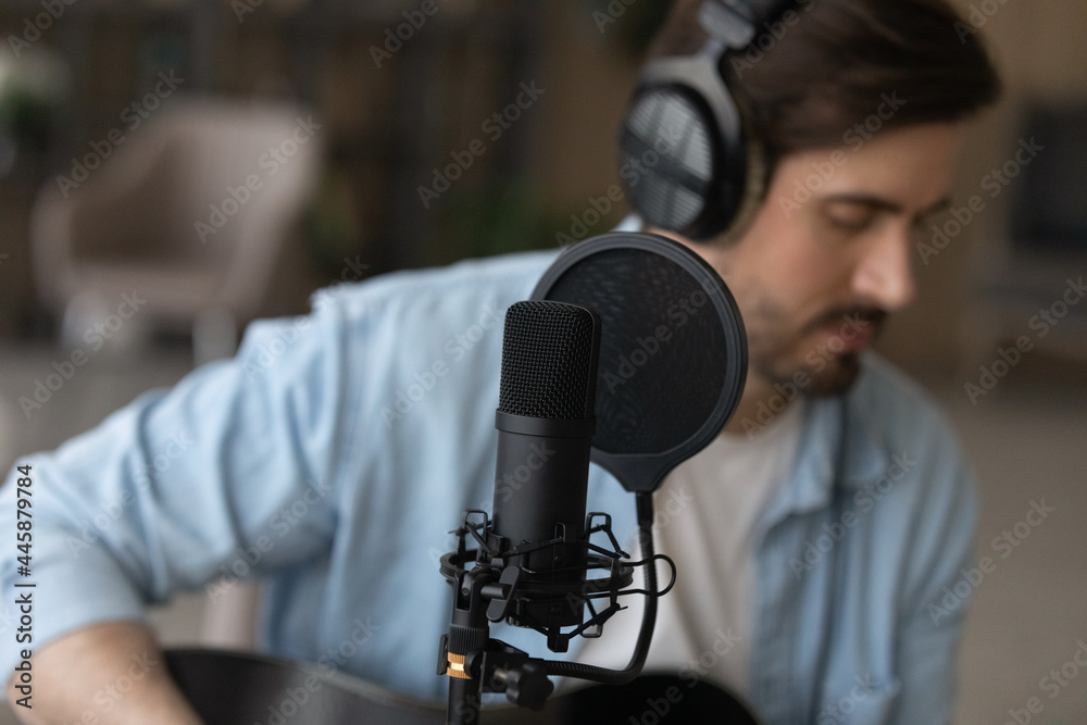 Close up focus on professional stand microphone with blurred young male musician singing on background. Young man in wired headphones recording own music, playing on guitar in modern domestic studio.