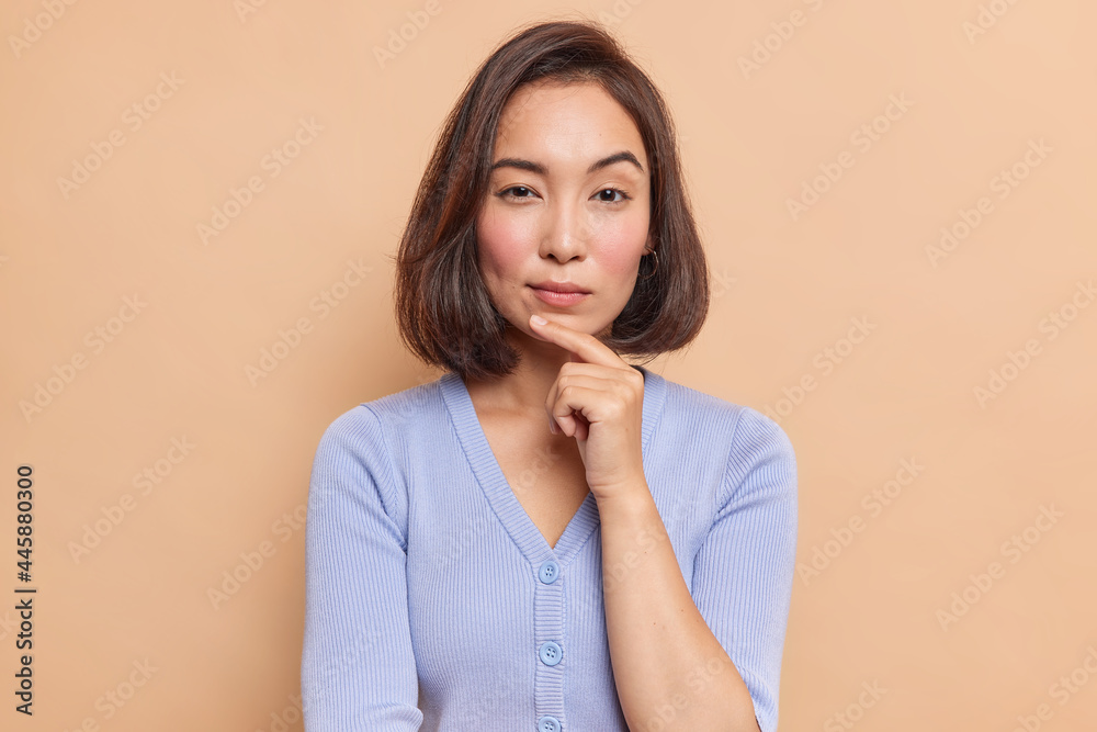 Portrait of serious dark haired Asian woman keeps finger on chin looks mysteriously at camera considers something dressed in blue jumper isolated over brown background. Let me think about it Portrait of serious dark haired Asian woman keeps finger on chin looks mysteriously at camera considers something dressed in blue jumper isolated over brown background. Let me think about it