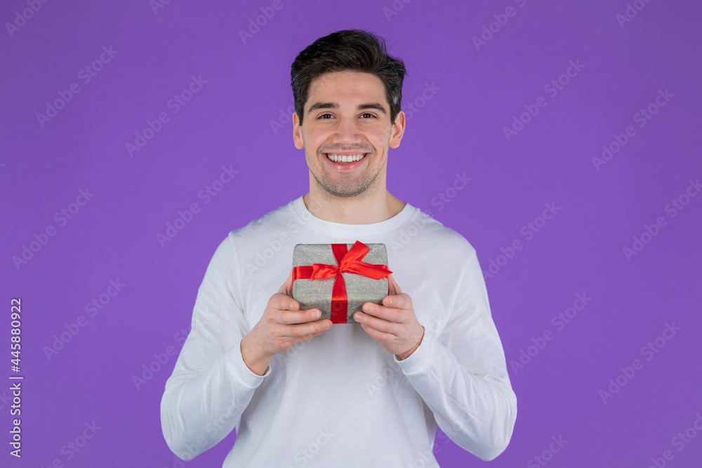 Handsome man holding gift box on purple studio background and smiles to camera. Happy european guy received present and interested in what's inside. Handsome man holding gift box on purple studio background and smiles to camera. Happy european guy received present and interested in what's inside.