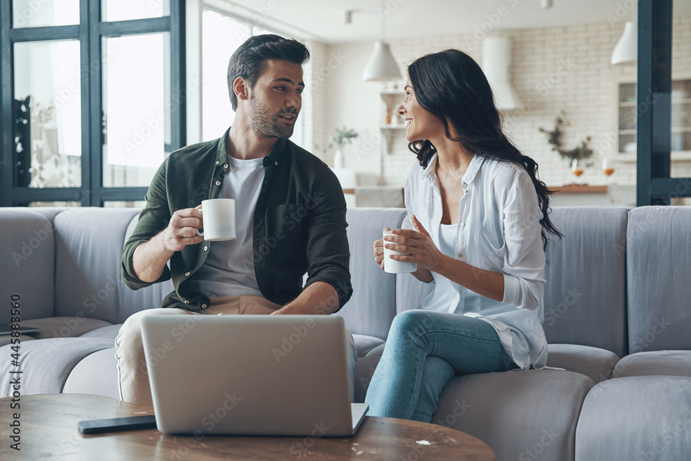 Beautiful young couple drinking coffee and looking on the laptop while sitting on the sofa at home Beautiful young couple drinking coffee and looking on the laptop while sitting on the sofa at home