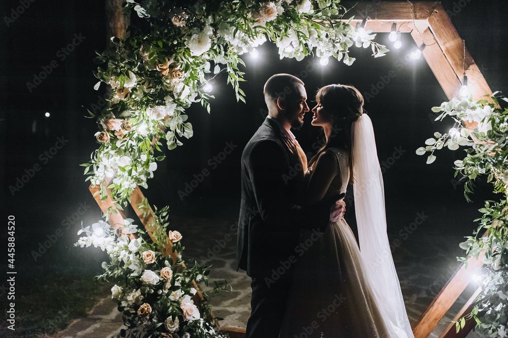 Φωτογραφία Stylish groom in a vest and a beautiful bride in a white long dress hug near a wooden arch, decorated with flowers, electric lamps, in nature in the forest at night