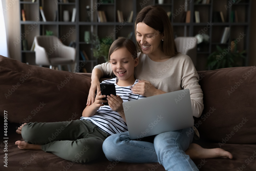 Happy gen Z girl and mom relaxing on couch with gadgets. Mother and little daughter kid using parental control app on smartphone, playing online game, watching video on laptop, browsing internet Happy gen Z girl and mom relaxing on couch with gadgets. Mother and little daughter kid using parental control app on smartphone, playing online game, watching video on laptop, browsing internet
