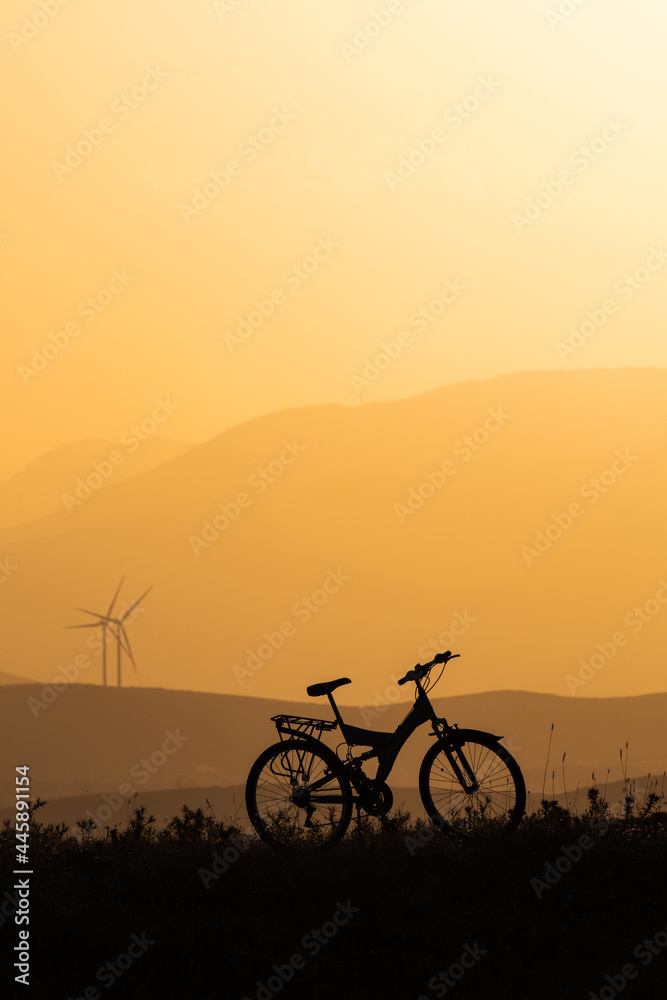 Fototapeta premium Mountains bike silhouette and setting sun, orang sunset, wind turbines and mountains on background.