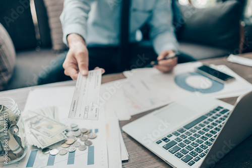 Фотография A Businessperson's hand giving cheque to customer and dollar bill, coin, laptop and graph chart on the desk at office