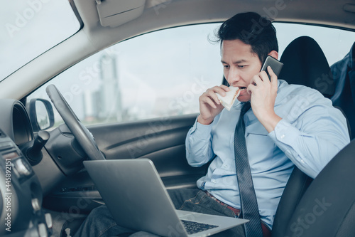 Businessman eating sandwich while working in laptop and talking on the phone in the drivers seat in his car. Busy businessman and food in car.