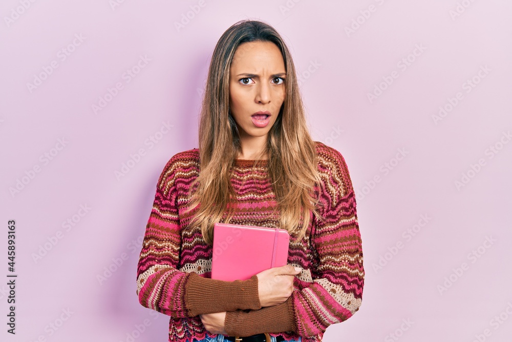 Beautiful hispanic woman hugging book in shock face, looking skeptical and sarcastic, surprised with open mouth Beautiful hispanic woman hugging book in shock face, looking skeptical and sarcastic, surprised with open mouth