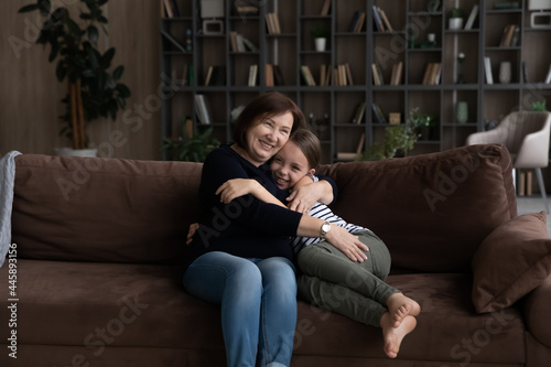 Happy grandma hugging and cuddling cute granddaughter on couch at home. Grandmother enjoying meeting and leisure with grandkid, expressing love and affection, intergenerational family relationship