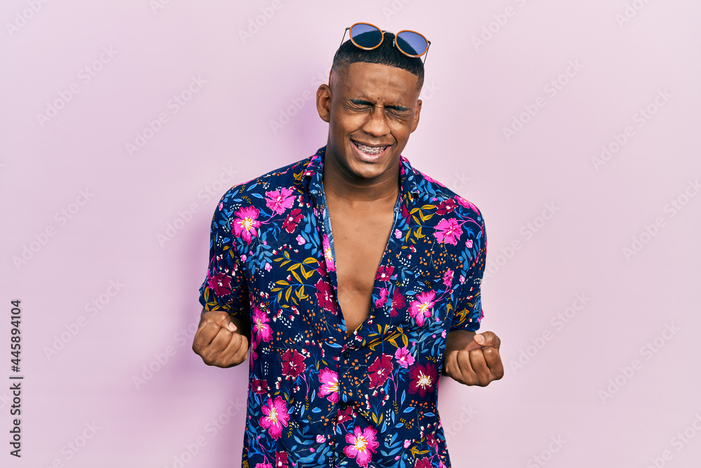 Fototapeta premium Young black man wearing hawaiian shirt and sunglasses very happy and excited doing winner gesture with arms raised, smiling and screaming for success. celebration concept.