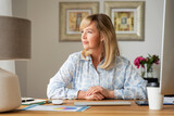 Blond haired attractive woman looking thoughtfully while sitting at desk at home and working