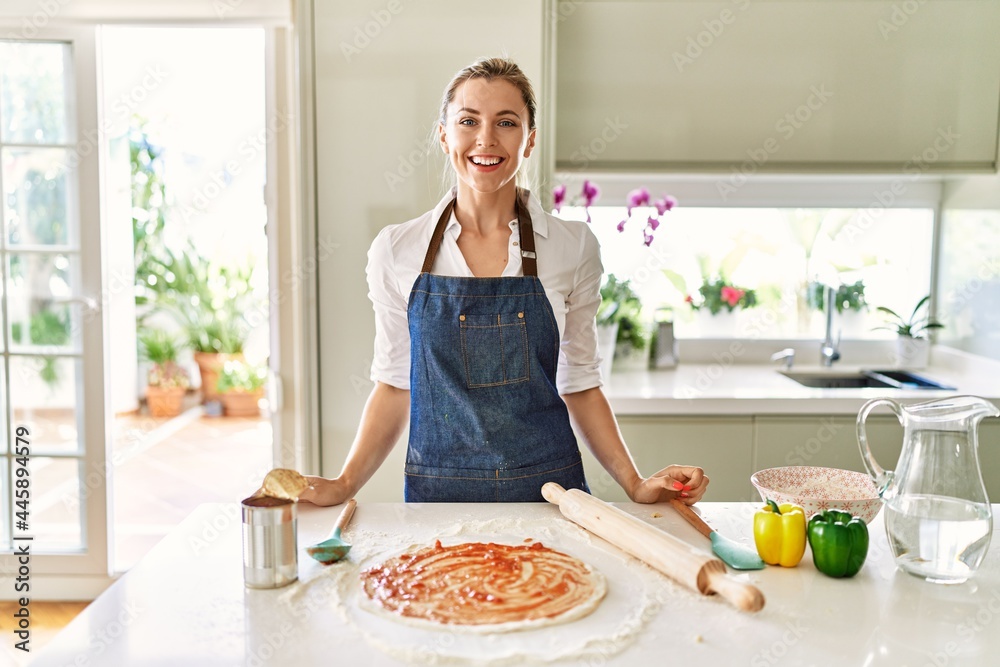 Beautiful blonde woman wearing apron cooking pizza looking positive and happy standing and smiling with a confident smile showing teeth