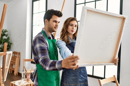 Two hispanic students with relaxed expression holding canvas at art school.