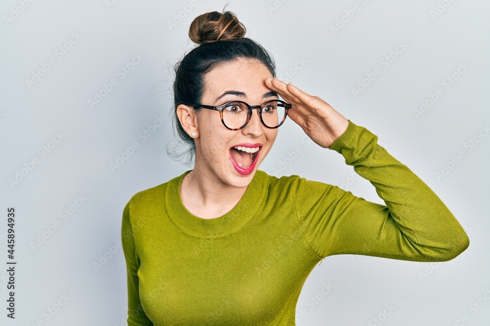 Young hispanic girl wearing casual clothes and glasses very happy and smiling looking far away with hand over head. searching concept.