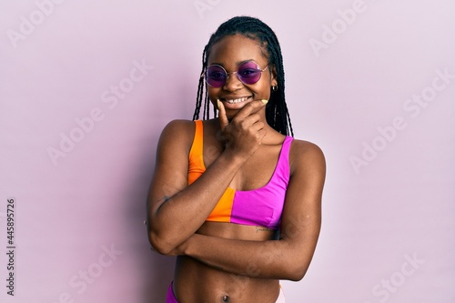Young african american woman wearing bikini and sunglasses looking confident at the camera smiling with crossed arms and hand raised on chin. thinking positive.