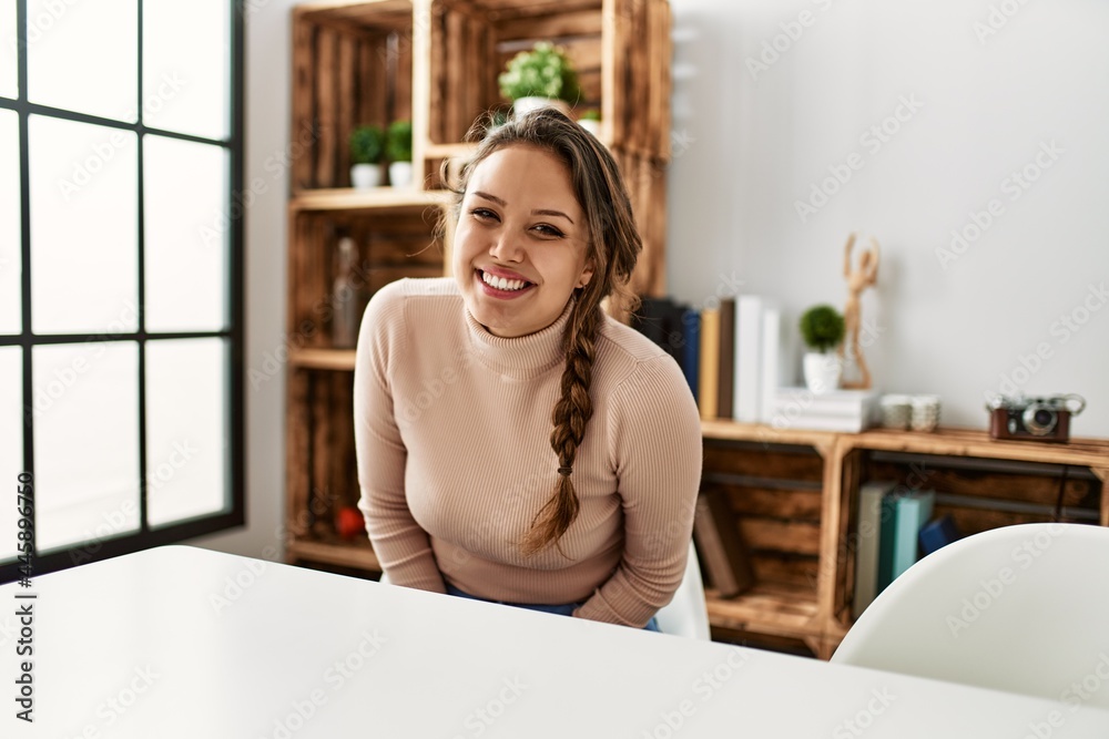 Young hispanic girl wearing casual clothes sitting on the table at home looking positive and happy standing and smiling with a confident smile showing teeth Young hispanic girl wearing casual clothes sitting on the table at home looking positive and happy standing and smiling with a confident smile showing teeth