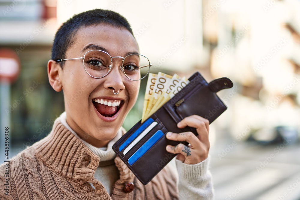 Beautiful hispanic woman with short hair smiling happy outdoors holding wallet with norwegian banknotes Beautiful hispanic woman with short hair smiling happy outdoors holding wallet with norwegian banknotes