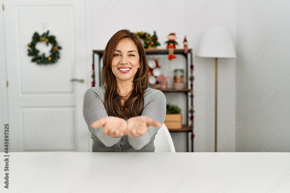 Young latin woman sitting on the table by christmas decor smiling with hands palms together receiving or giving gesture. hold and protection Young latin woman sitting on the table by christmas decor smiling with hands palms together receiving or giving gesture. hold and protection