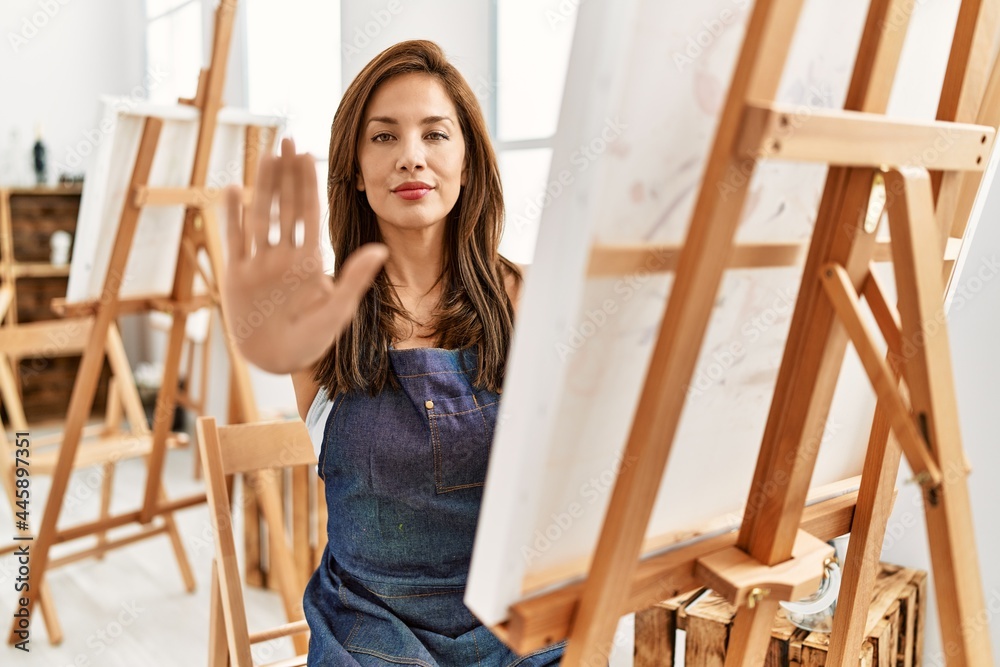 Young latin artist woman painting at art studio with open hand doing stop sign with serious and confident expression, defense gesture