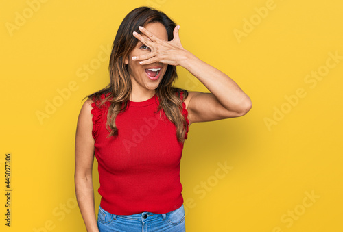 Young hispanic woman wearing casual clothes peeking in shock covering face and eyes with hand, looking through fingers with embarrassed expression.