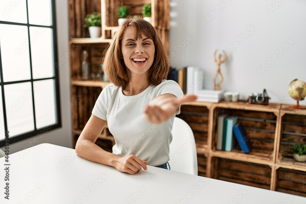 Young caucasian girl wearing casual clothes sitting on the table at home smiling friendly offering handshake as greeting and welcoming. successful business. Young caucasian girl wearing casual clothes sitting on the table at home smiling friendly offering handshake as greeting and welcoming. successful business.