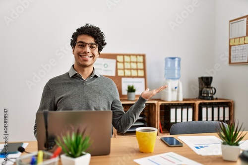 Young hispanic man wearing business style sitting on desk at office smiling cheerful presenting and pointing with palm of hand looking at the camera.