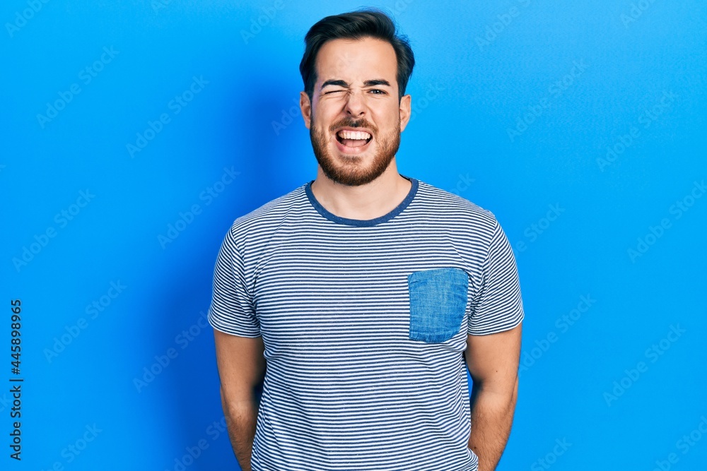 Handsome caucasian man with beard wearing casual striped t shirt winking looking at the camera with sexy expression, cheerful and happy face. Handsome caucasian man with beard wearing casual striped t shirt winking looking at the camera with sexy expression, cheerful and happy face.