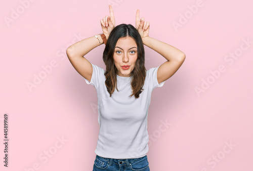 Young beautiful woman wearing casual white t shirt doing funny gesture with finger over head as bull horns