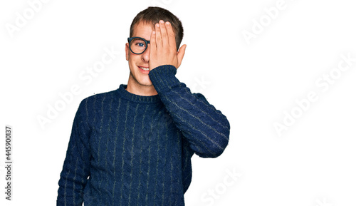 Young blond man wearing casual clothes and glasses covering one eye with hand, confident smile on face and surprise emotion.