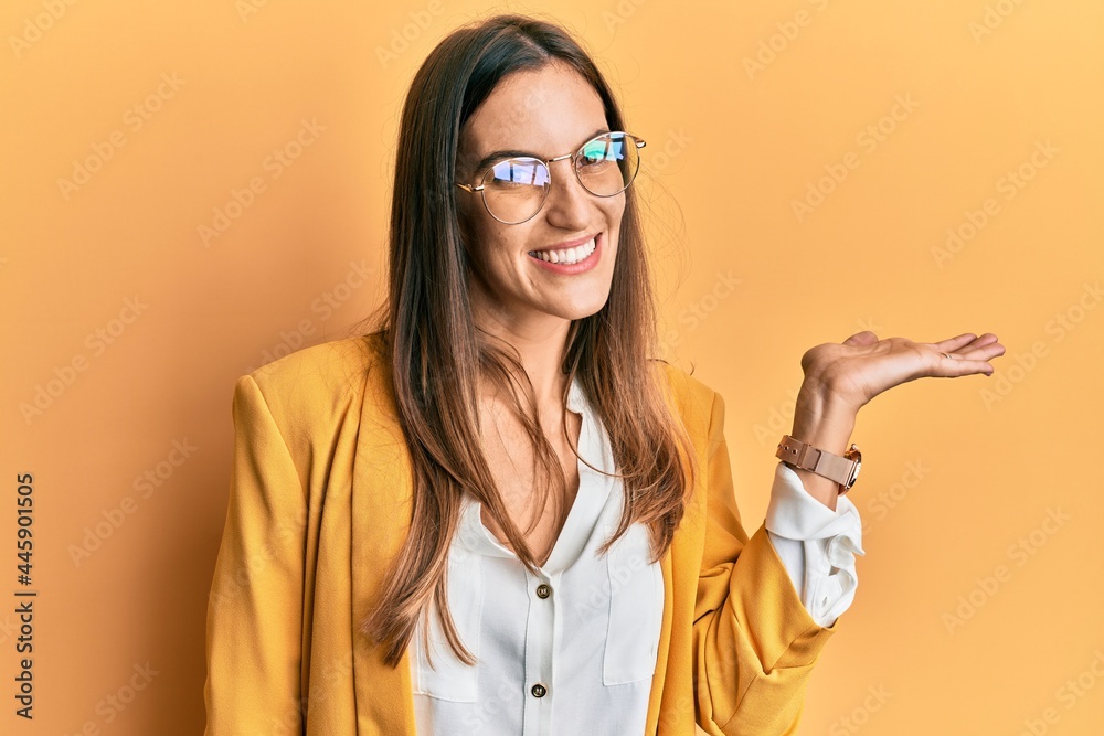 Young beautiful woman wearing business style and glasses smiling cheerful presenting and pointing with palm of hand looking at the camera.