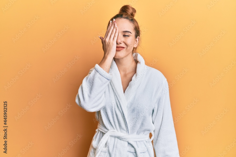 Young blonde woman wearing bathrobe covering one eye with hand, confident smile on face and surprise emotion. Young blonde woman wearing bathrobe covering one eye with hand, confident smile on face and surprise emotion.