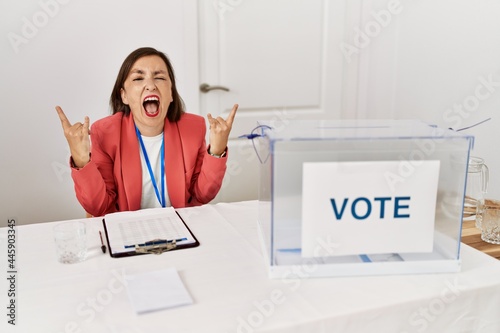 Beautiful middle age hispanic woman at political election sitting by ballot shouting with crazy expression doing rock symbol with hands up. music star. heavy music concept.