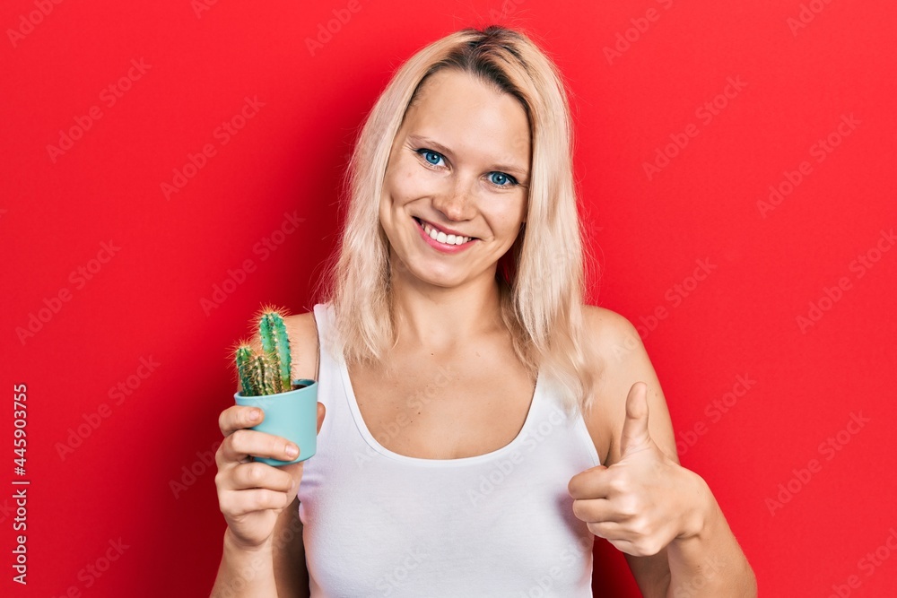 Beautiful caucasian blonde woman holding small cactus pot smiling happy and positive, thumb up doing excellent and approval sign Beautiful caucasian blonde woman holding small cactus pot smiling happy and positive, thumb up doing excellent and approval sign