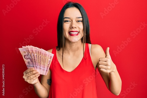 Young hispanic girl holding thai baht banknotes smiling happy and positive, thumb up doing excellent and approval sign