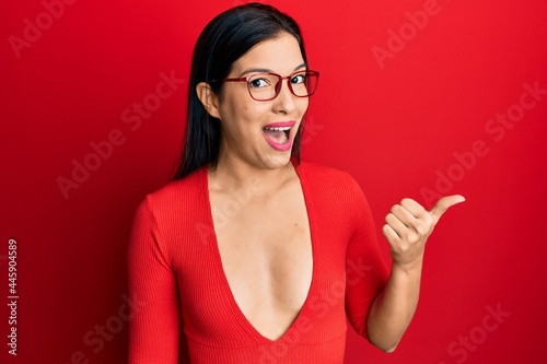 Young latin woman wearing casual clothes and glasses smiling with happy face looking and pointing to the side with thumb up.