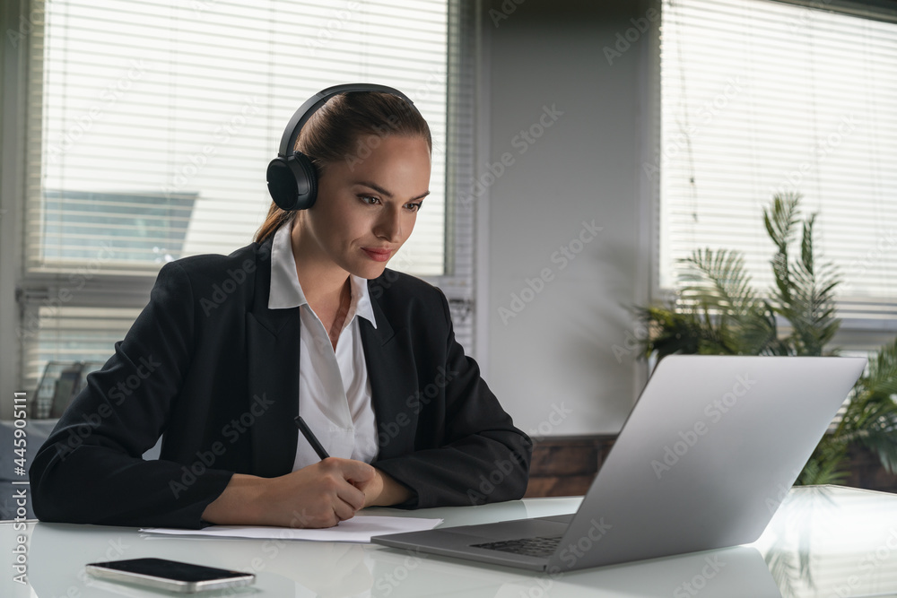 Portrait of thoughtful attractive business woman in suit sitting at the desk in front of laptop and dreaming about career opportunities. Taking notes and listening to podcast. Office background. Portrait of thoughtful attractive business woman in suit sitting at the desk in front of laptop and dreaming about career opportunities. Taking notes and listening to podcast. Office background.