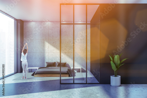 Quadro su tela Rear view of just woke up woman who is holds her hands up in front of panoramic window with city view, cozy home wear, modern panoramic studio apartment with bed