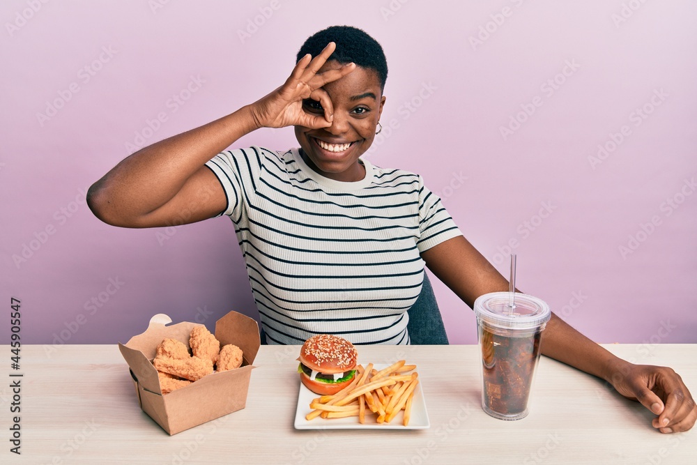 Young african american woman eating a tasty classic burger with fries and soda smiling happy doing ok sign with hand on eye looking through fingers Young african american woman eating a tasty classic burger with fries and soda smiling happy doing ok sign with hand on eye looking through fingers