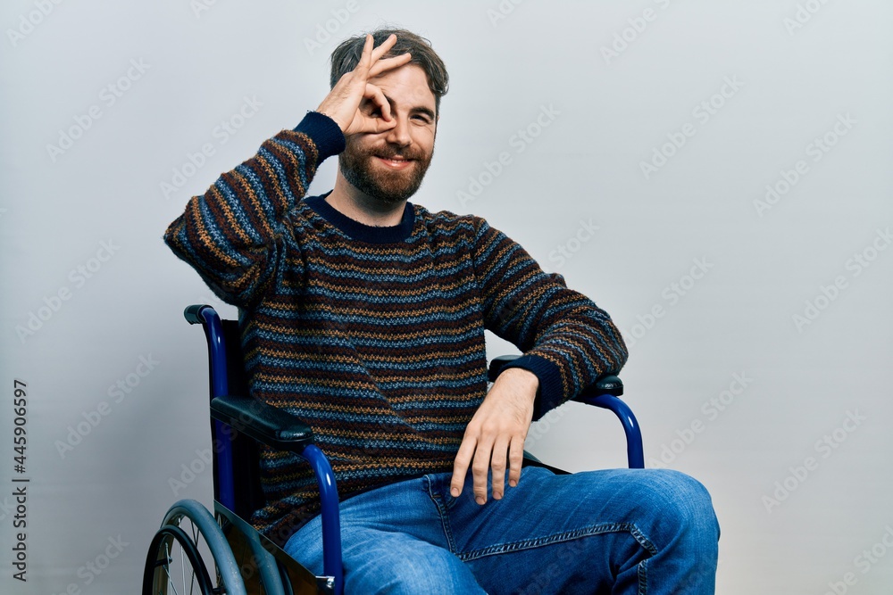 Caucasian man with beard sitting on wheelchair smiling happy doing ok sign with hand on eye looking through fingers Caucasian man with beard sitting on wheelchair smiling happy doing ok sign with hand on eye looking through fingers