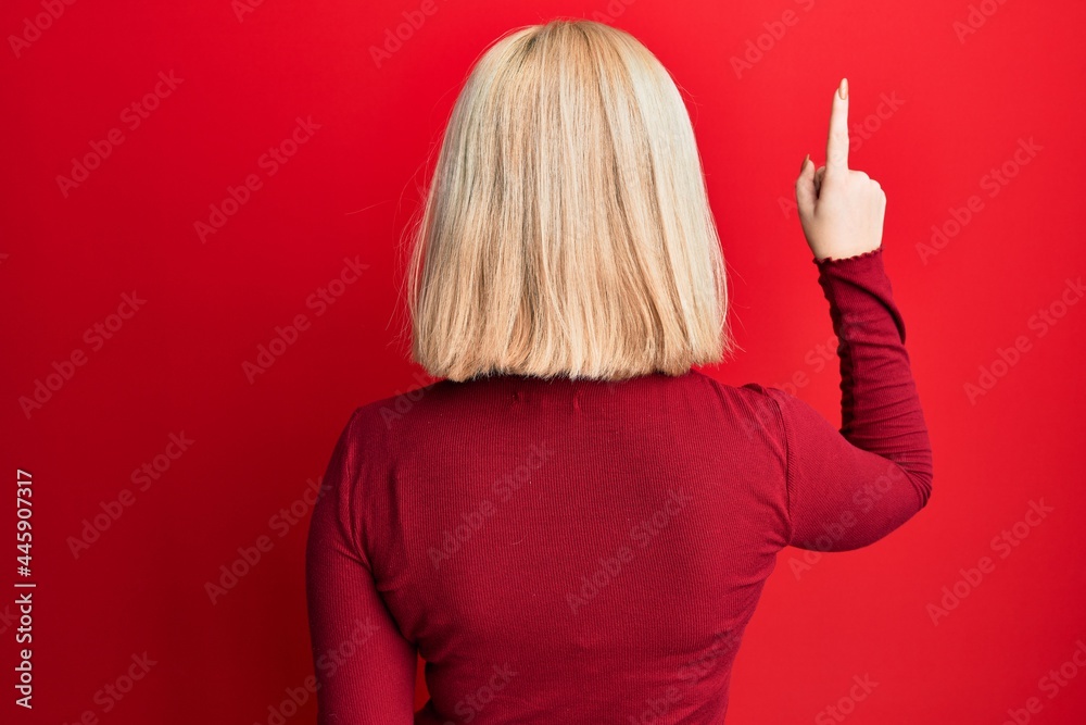 Young blonde woman wearing casual clothes posing backwards pointing ahead with finger hand Young blonde woman wearing casual clothes posing backwards pointing ahead with finger hand