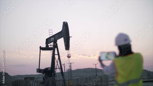 Construction worker surveys oil rig with device at sunrise. 